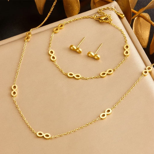 Gold jewelry set featuring a necklace, bracelet, and stud earrings with infinity symbol charms, displayed on a beige jewelry box.