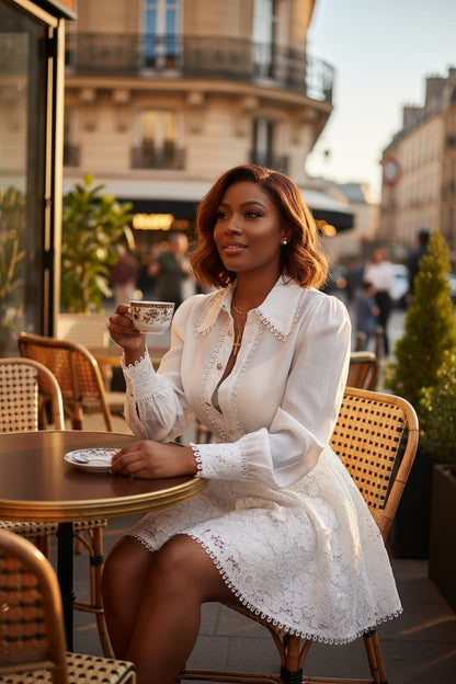 A woman sits at a cafe table wearing a white lace long-sleeve dress, holding a teacup and saucer in a Parisian-style outdoor setting.