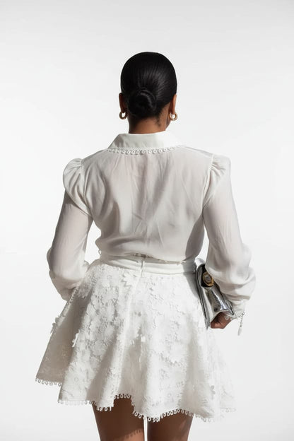 A woman shown from behind wearing a white long-sleeve blouse with puffed sleeves and a matching white textured mini skirt, holding a metallic clutch against a plain white studio background.